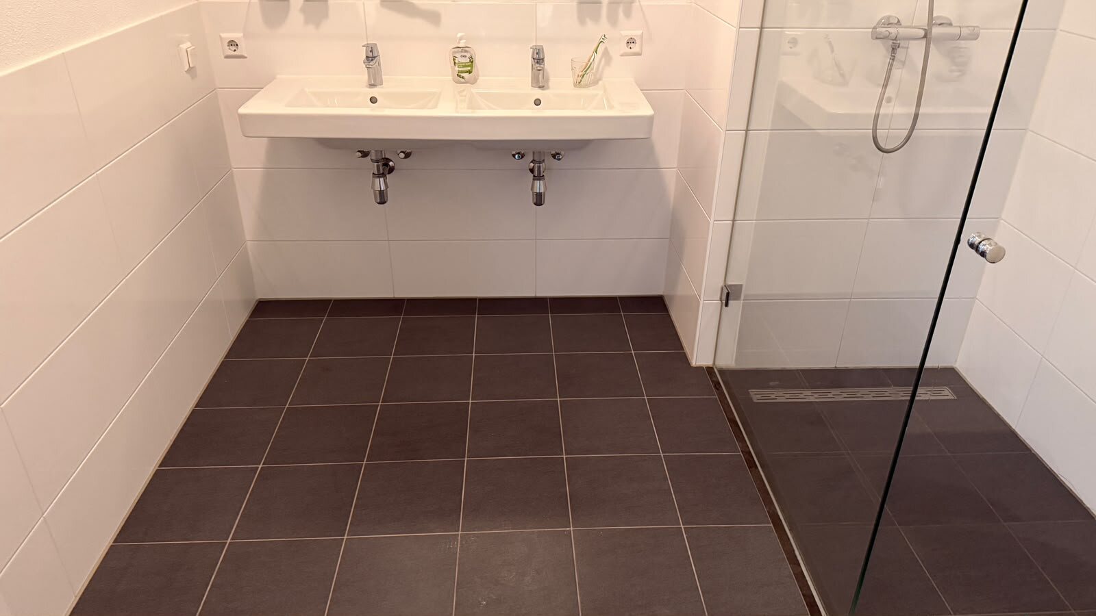 Badkamer na de deep clean: dubbele wastafel, donkere vloertegels en een heldere glazen douchedeur met knop